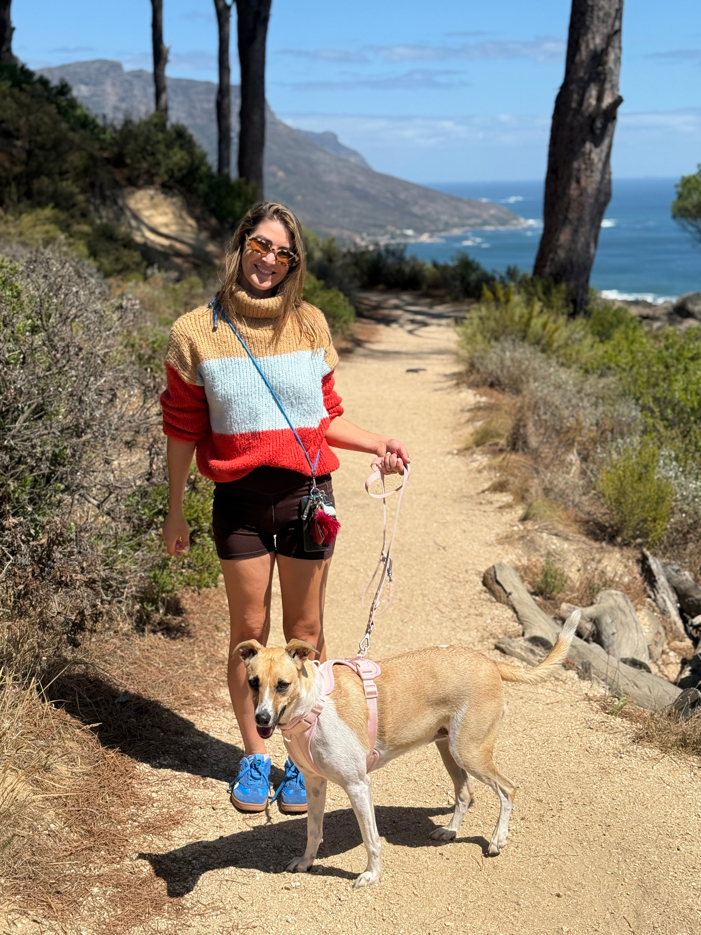 Woman in a colourful striped jumper walking a tan dog on a coastal mountain trail with the sea behind